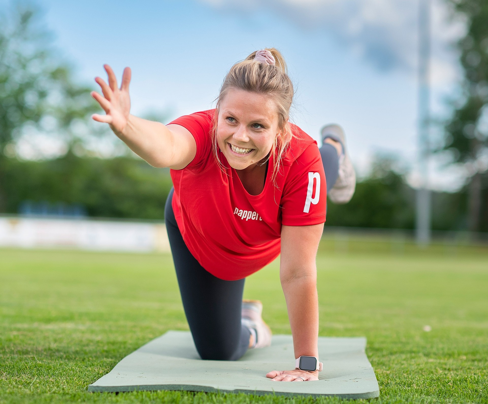 Fitnessbäcker Kurs-Teilnehmerin in rotem Pappert-T-Shirt macht Yoga-Übungen auf einer Matte im Freien und lächelt dabei in die Kamera.